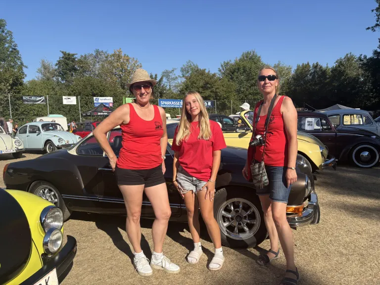 Three women in red t-shirts and shorts stand in front of a black car, smiling and posing for a photo at a car show.