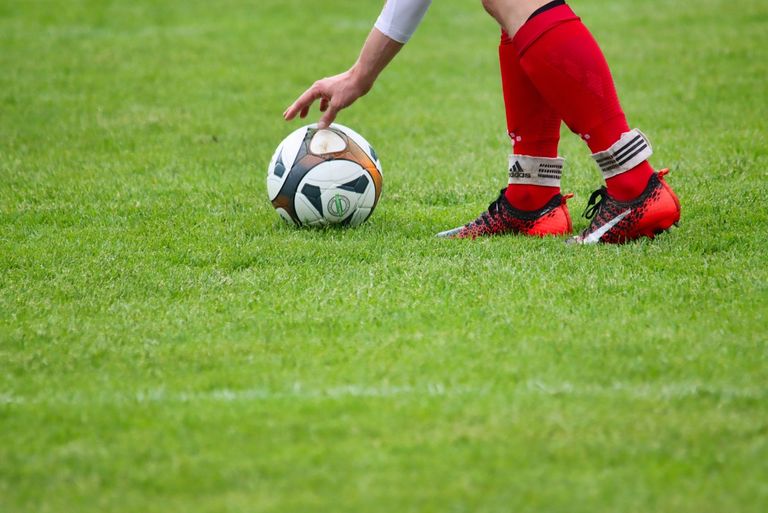 Ein Fußballspieler in roten Shorts und Adidas-Schuhen berührt einen Fußball auf einem grünen Feld.