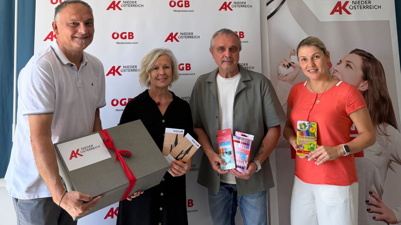 Four people stand in front of a banner with AK Niederosterreich logos, holding gift boxes and packs of colored pencils. The woman on the left holds a large box with a red ribbon.