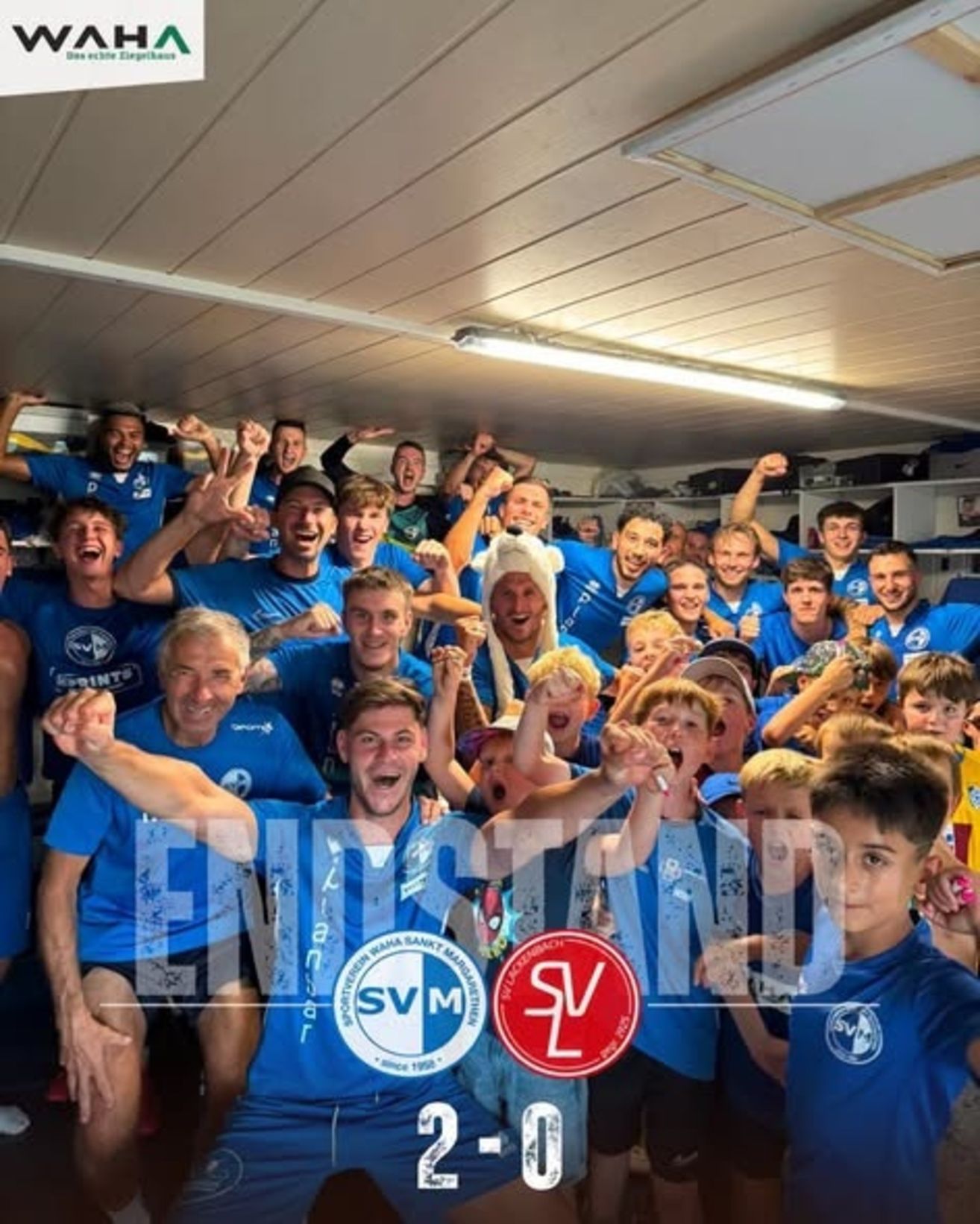 Eine Gruppe von Menschen in blauen Trikots feiert in einem Raum. Sie heben ihre Hände und einige tragen Hüte. Der Raum hat weiße Deckenpaneele und Regale mit Gegenständen darauf. In der Mitte des Bildes befindet sich ein Logo.