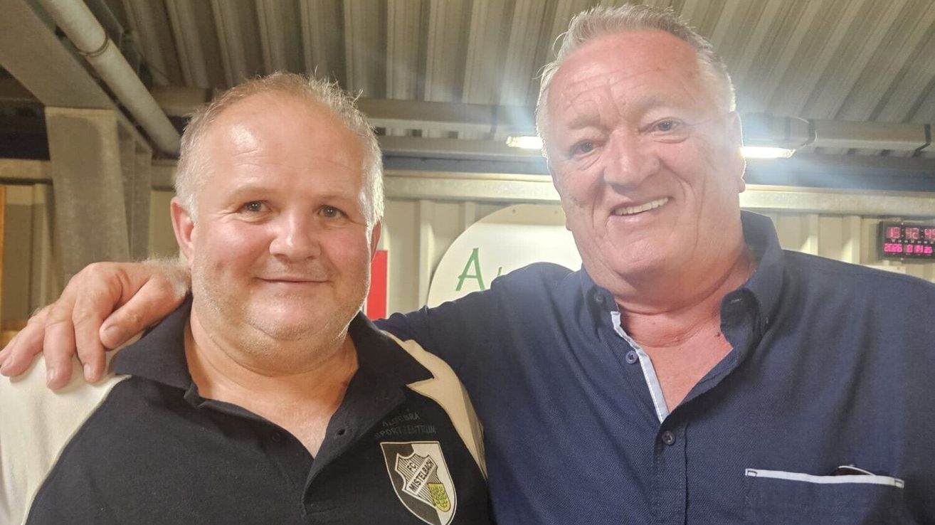 Two men are smiling and posing for a photo. The man on the left is wearing a black shirt with a logo. The man on the right has his arm around the other man's shoulder.