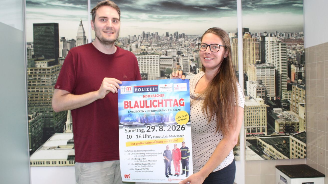 A man and a woman are standing in front of a poster for the Blaulichttag event, holding it up for the camera. The poster features a cityscape in the background and includes details about the event date and activities.