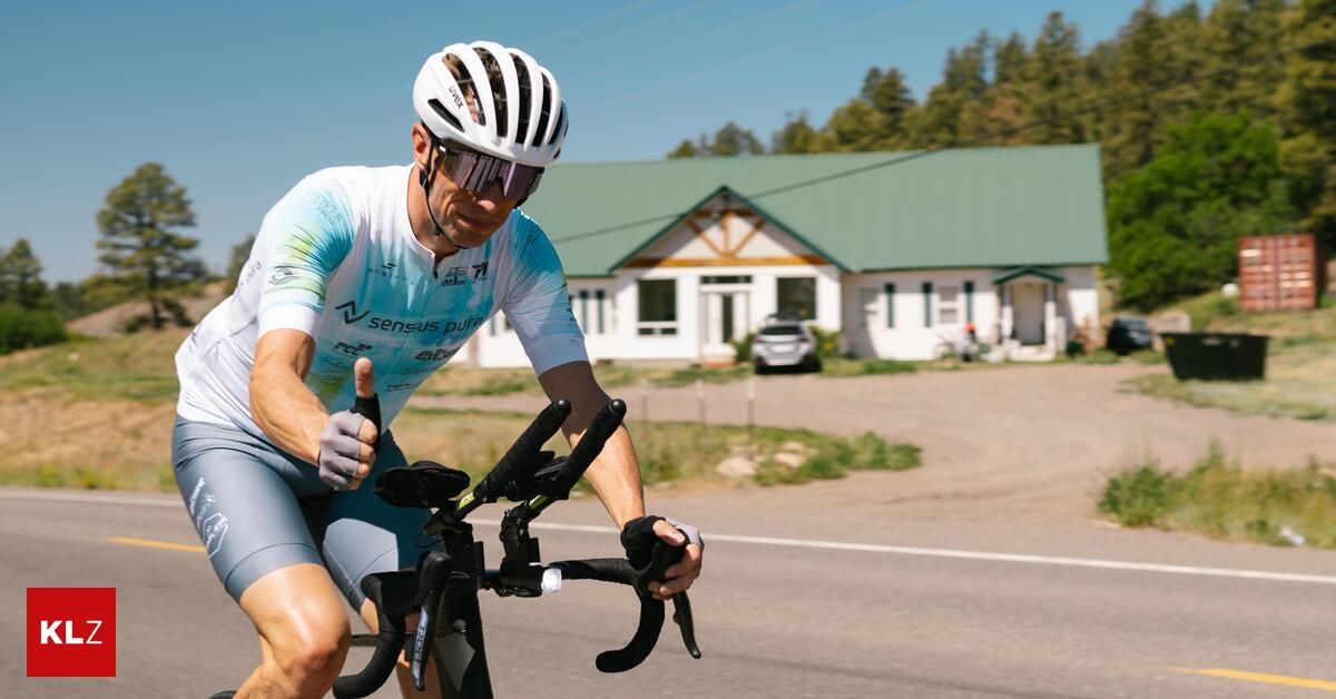 A cyclist wearing a white helmet and cycling jersey rides down a road with a building in the background.