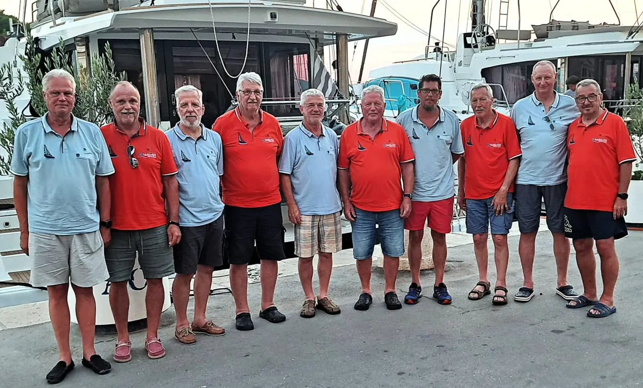 Six older men in matching red and blue shirts pose for a photo in front of a boat.