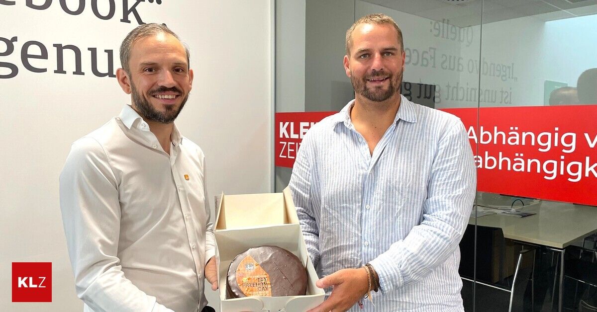Two men in a modern office setting, one holding a box containing a chocolate cake, both smiling and posing for a photo.