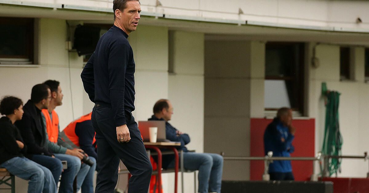 A man in a dark blue long-sleeve shirt and pants walks across a field, possibly a coach, with people sitting in the background.