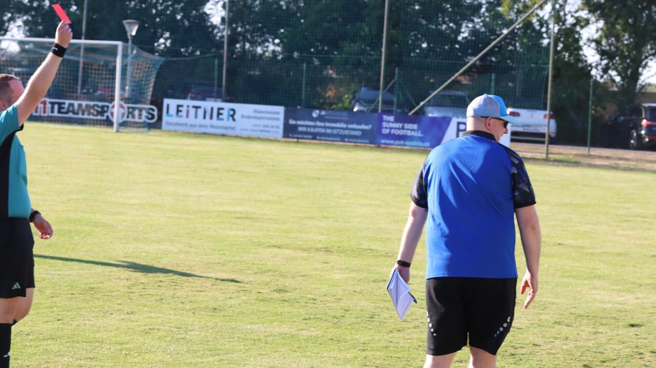 A man in a blue shirt and black shorts is standing on a grassy field, holding a notebook and a pen, possibly a coach or referee.