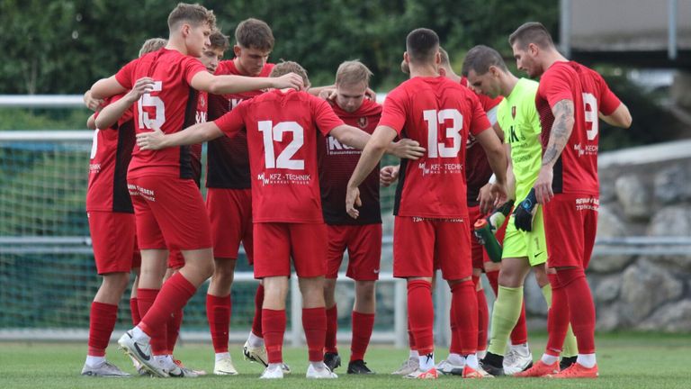 A group of soccer players in red uniforms are huddled together on the field, likely discussing their strategy. The player in the center wears the number 12, while the player next to him wears the number 13.