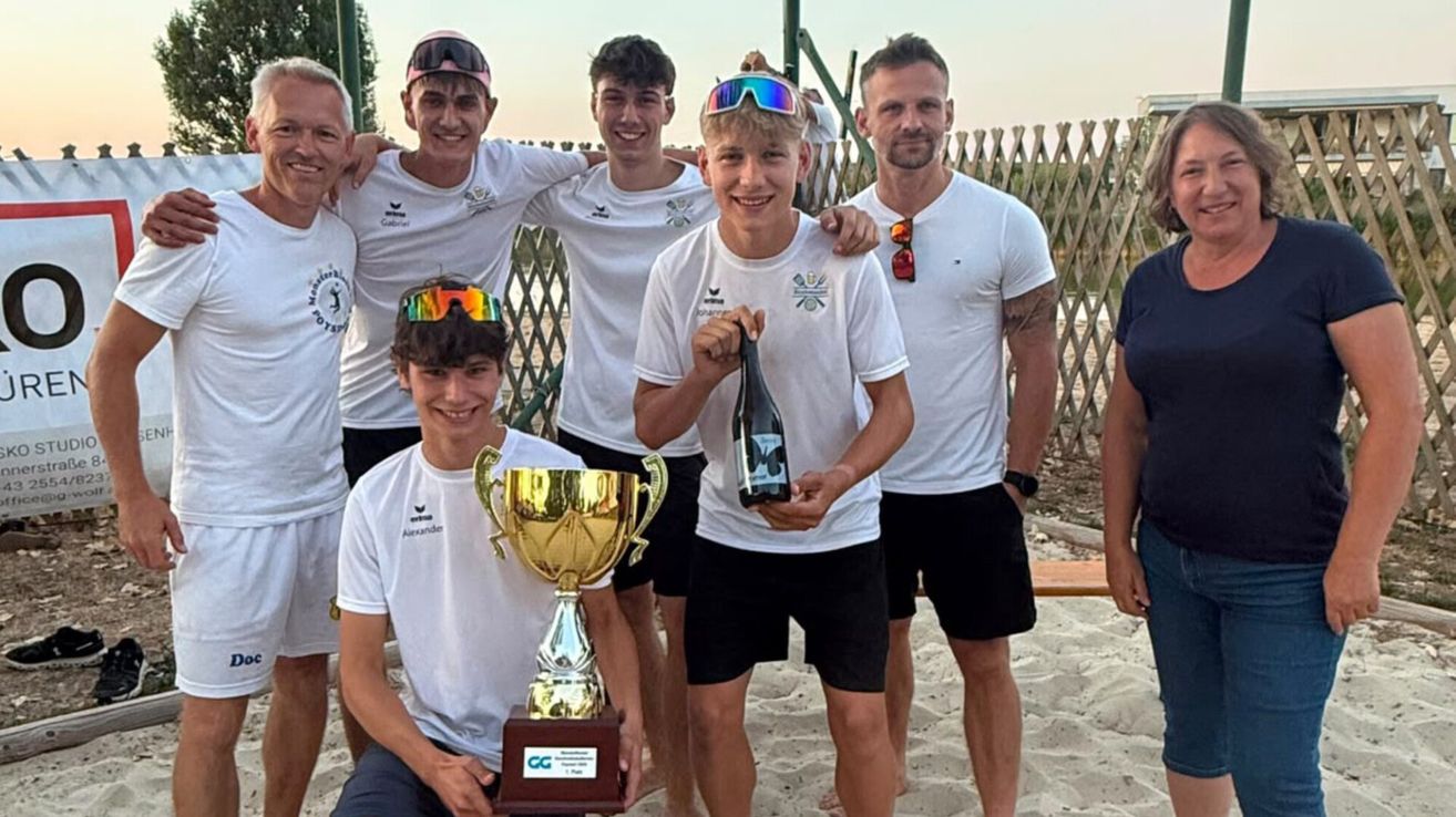 Eine Gruppe junger Männer in weißen Shirts und schwarzen Shorts posiert für ein Foto am Strand, einer hält einen Pokal und der andere eine Flasche.
