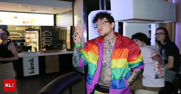 A man wearing a colorful rainbow jacket and glasses poses for a photo in a public space.