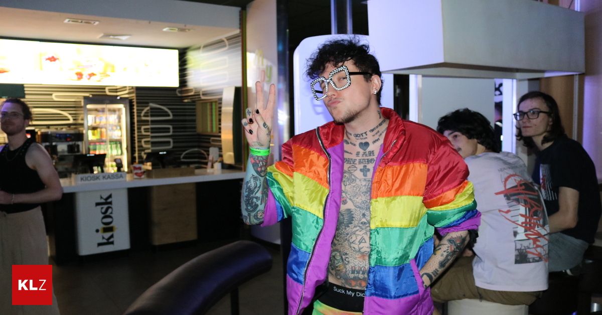 A man wearing a colorful rainbow jacket and glasses poses for a photo in a public space.