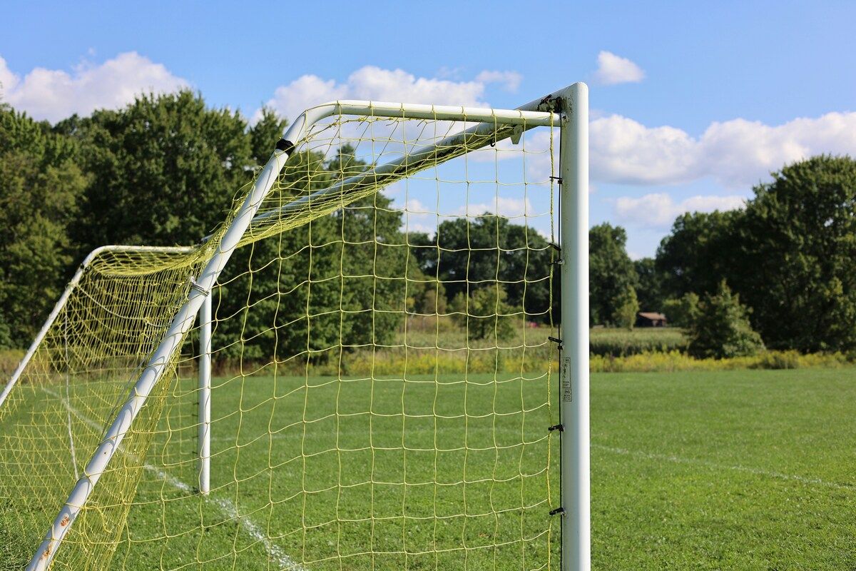 A soccer goal with a yellow net stands on a grassy field with trees in the background under a blue sky with clouds.