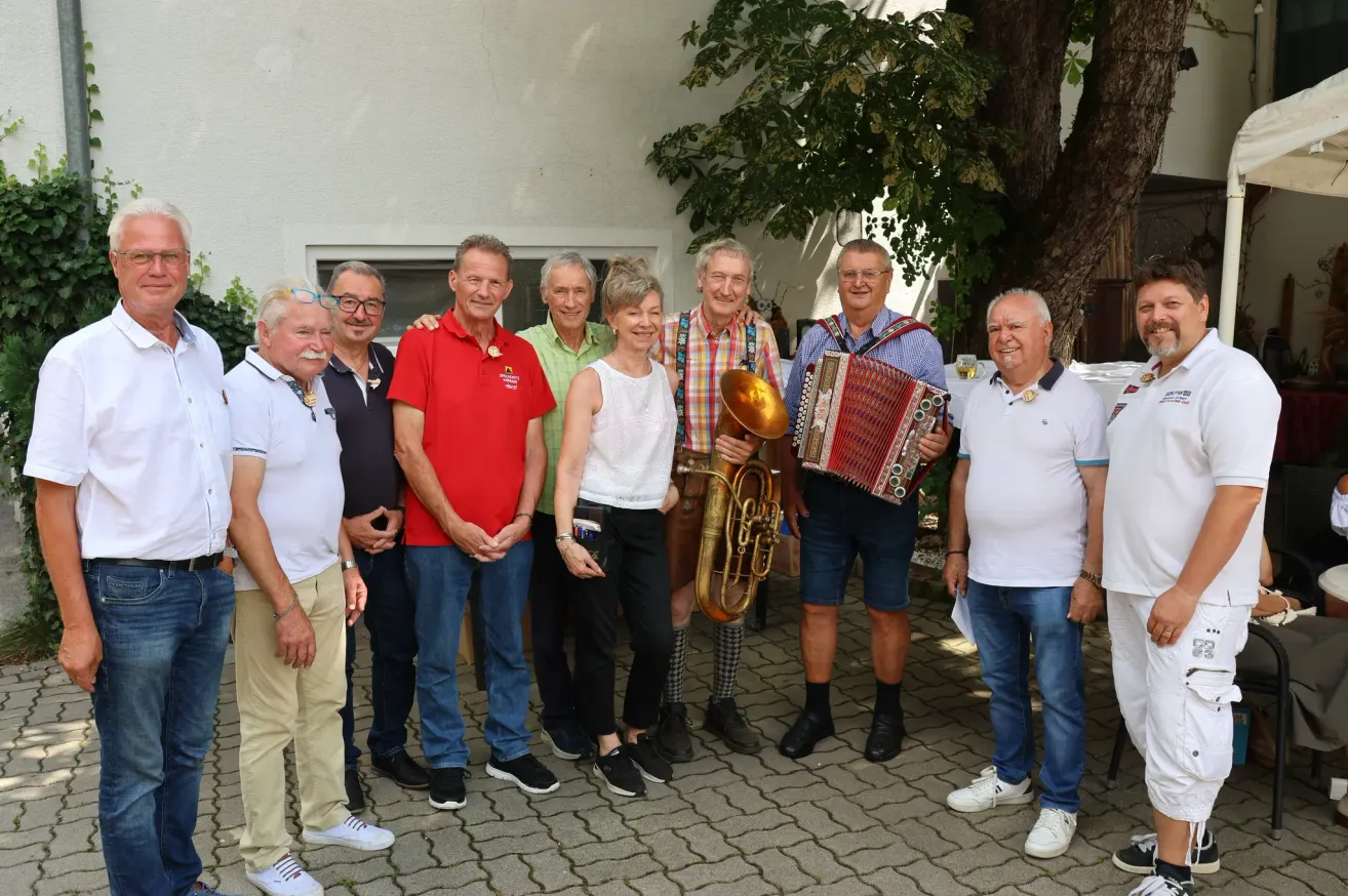 A group of older men and women are standing together in front of a white building, smiling for a photo. One man is holding a brass instrument, and another is playing an accordion.