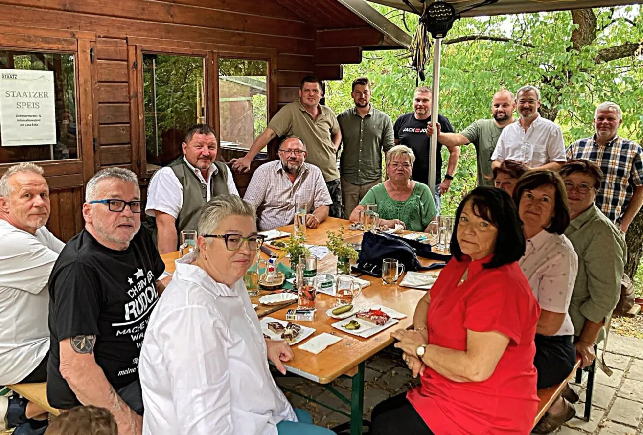A group of people, both men and women, are sitting around a table in a garden, smiling and posing for a photo. They are dressed casually.