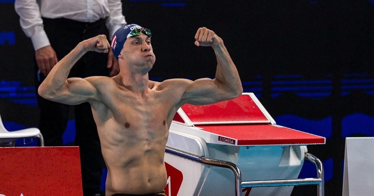 A swimmer celebrates with flexed arms and goggles on his head at a swimming event. He is wearing a blue swim cap with the word 'one' on it.