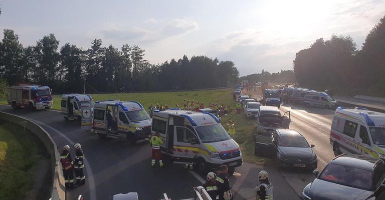 Emergency vehicles are parked on the side of a road, with several people in reflective vests attending to a situation. The vehicles include ambulances and fire trucks. The road is lined with trees and grass on both sides.