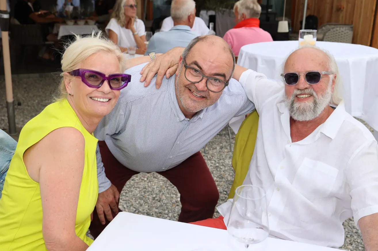 Three older adults smiling for a photo, one man with his arm around two women, at an outdoor event.