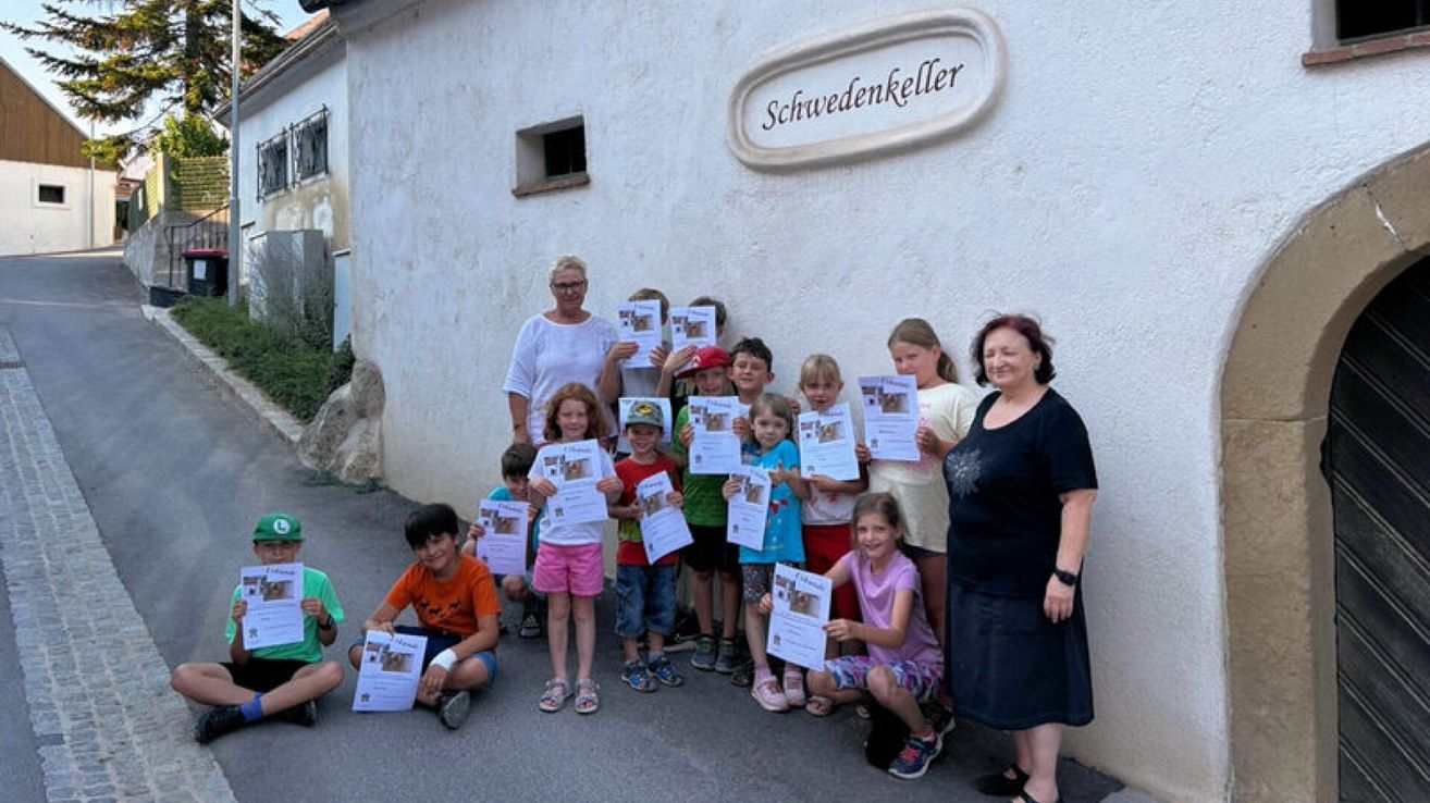 A group of children and adults stand in front of a building with a sign reading Schwedenkeller, holding certificates and smiling for a photo.