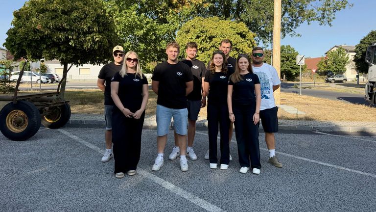 A group of people in matching black t-shirts and sneakers stand in a parking lot with trees in the background.