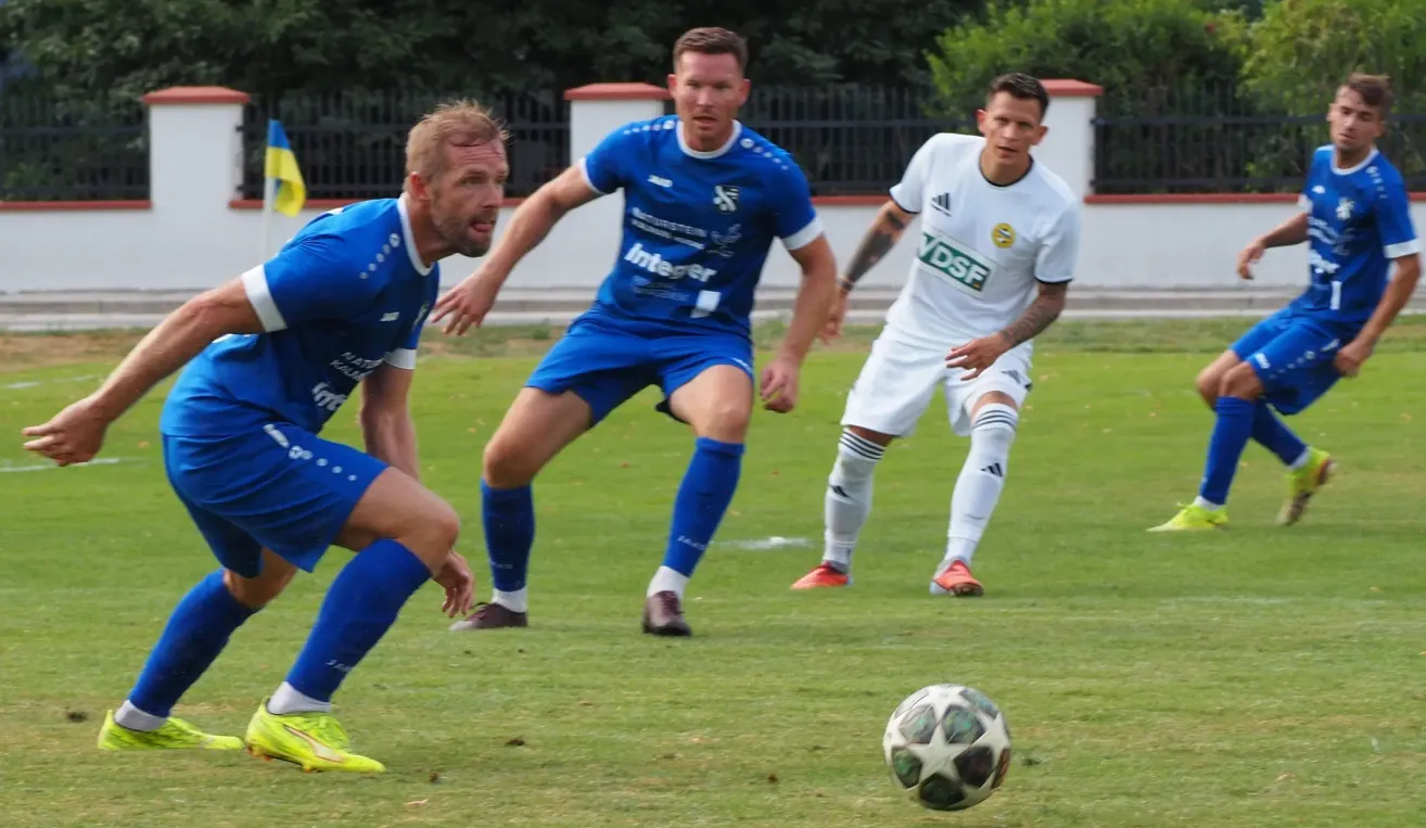 Three soccer players on a field, one in blue and two in white. The player in blue is looking at the ball while the other two are looking at him.