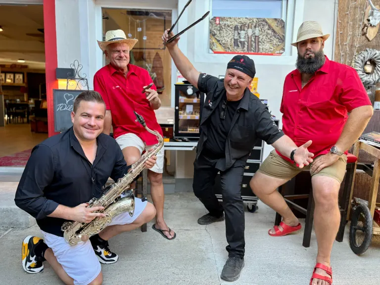 Four men in red shirts and hats, one holding a saxophone, another holding tongs, and two smiling at the camera. They are in a room with a glass door and a blackboard.