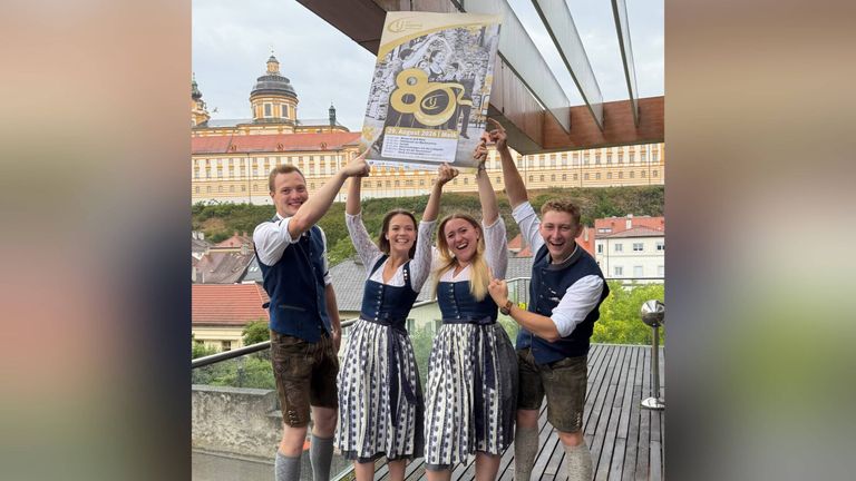 Four people in traditional Bavarian attire are holding up a poster with the number 80 on it in front of a historic building.