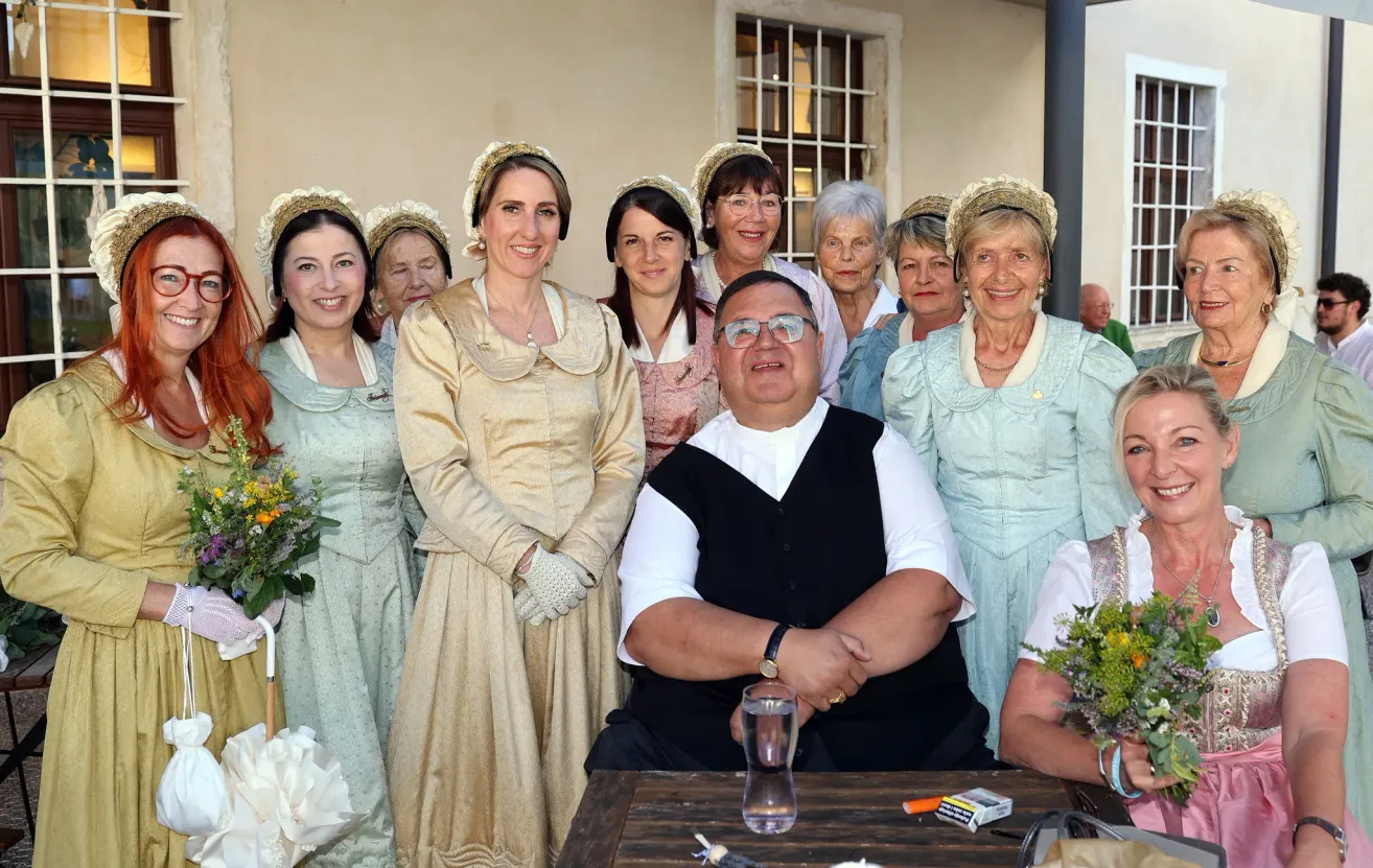 A group of women in vintage dresses and a man in a black vest pose for a photo outside. They are smiling and standing in front of a building.
