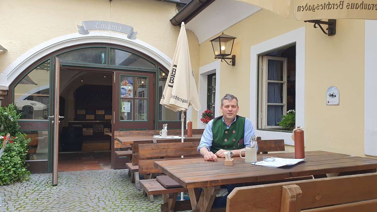A man in a green vest sits at a table in a restaurant with an umbrella and a mug.