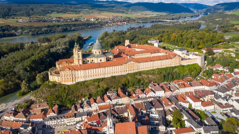 Aerial view of a large historical building with red roofs, surrounded by a town with many buildings and a river in the distance.