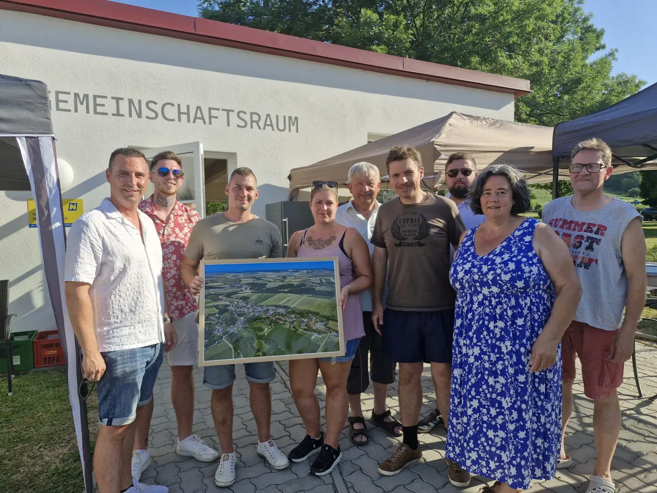 A group of people are standing outside a building with a sign that reads 'Meinschaftsraum'. They are holding a framed map and posing for a photo.