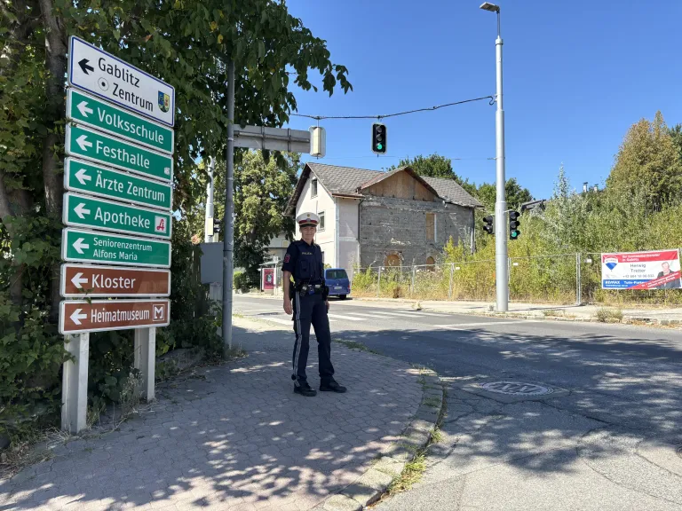 A police officer stands on a sidewalk with street signs pointing to various locations. The signs include directions to Gablitz Zentrum, Volksschule, Festhalle, Arzte Zentrum, Apotheke, Seniorenzentrum, Kloster, and Heimatmuseum. Behind the officer is a house with a fence and a blue van parked nearby.