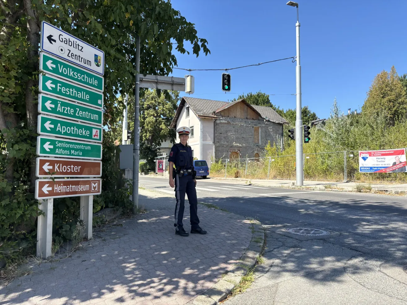 A police officer stands on a sidewalk with street signs pointing to various locations. The signs include directions to Gablitz Zentrum, Volksschule, Festhalle, Arzte Zentrum, Apotheke, Seniorenzentrum, Kloster, and Heimatmuseum. Behind the officer is a house with a fence and a blue van parked nearby.