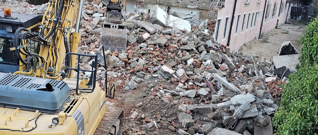 A yellow excavator is clearing debris from a demolished building, with a pile of rubble in front and a partially demolished building in the background.