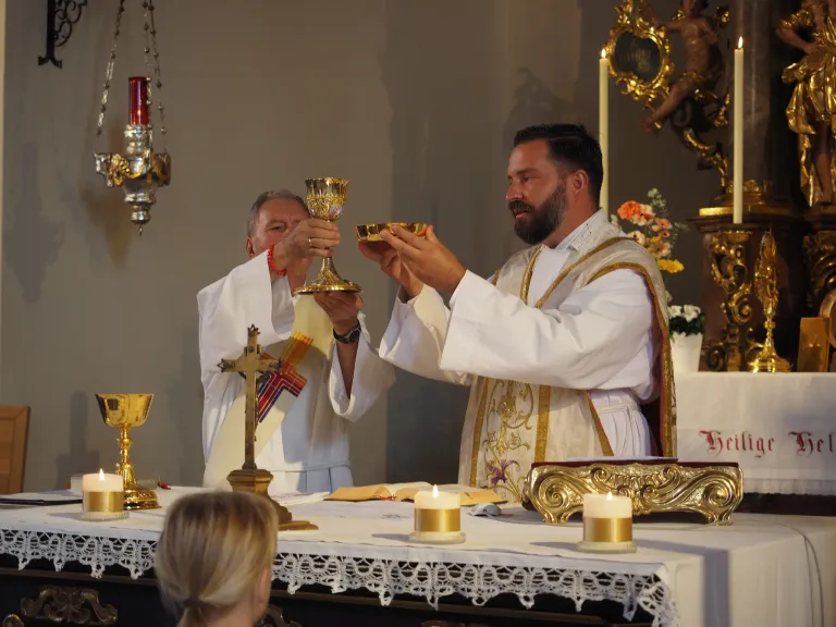 Zwei Priester halten goldene Kelche während einer religiösen Zeremonie in einer Kirche, mit Kerzen und einem Kreuz auf dem Altar.
