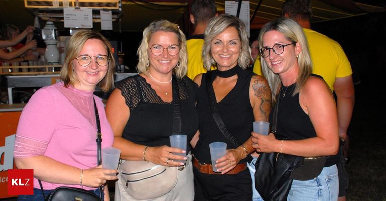 Four women are smiling and posing for a photo, holding plastic cups. They are dressed casually and appear to be at an outdoor event.