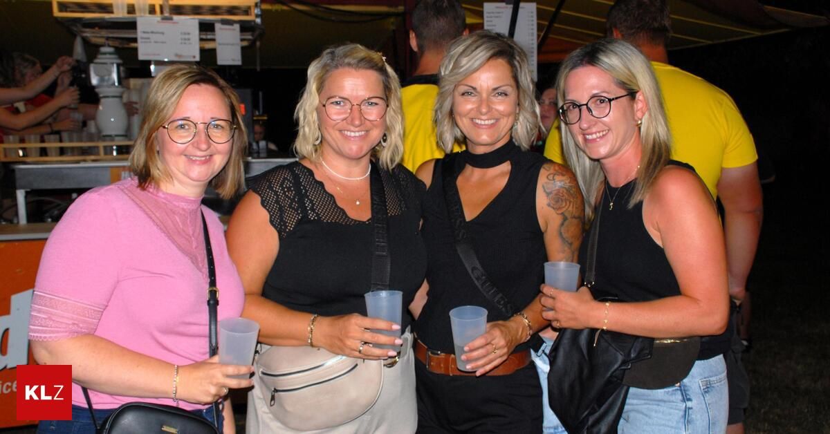Four women are smiling and posing for a photo, holding plastic cups. They are dressed casually and appear to be at an outdoor event.