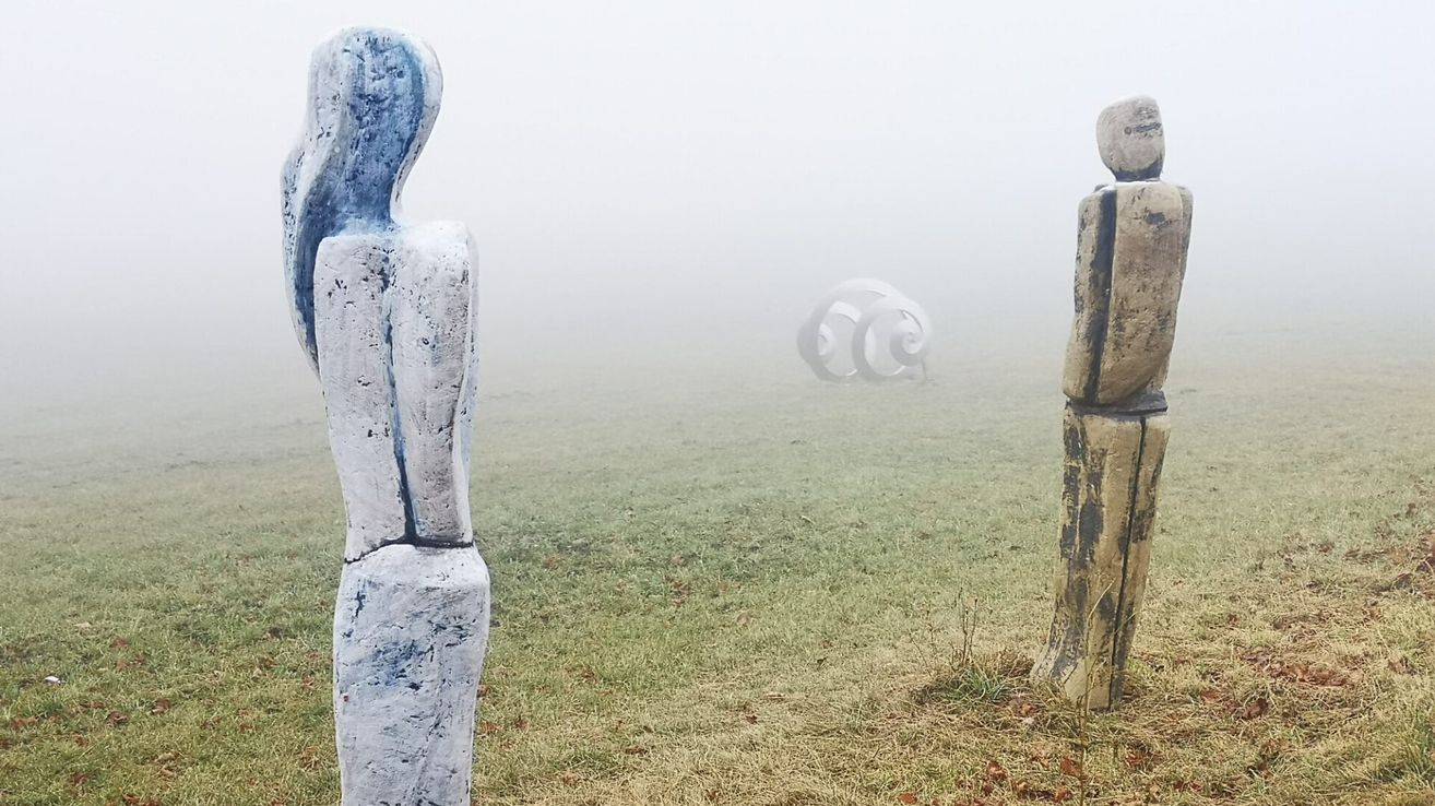 A foggy landscape with two abstract sculptures, one blue and white, the other brown, and a spiral object in the distance.