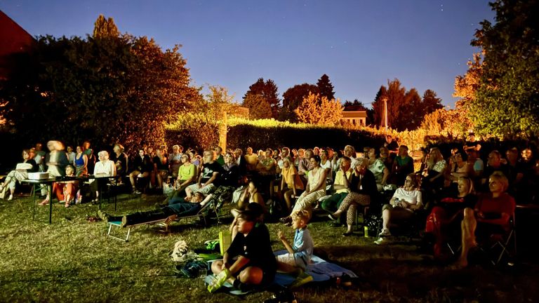 A large group of people are gathered outdoors in a grassy area for an evening event, some sitting on chairs and blankets, others standing, with trees and a building in the background.