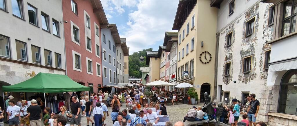 A bustling street market in a European town, with people shopping under umbrellas and tents, and buildings with intricate designs and a clock tower.