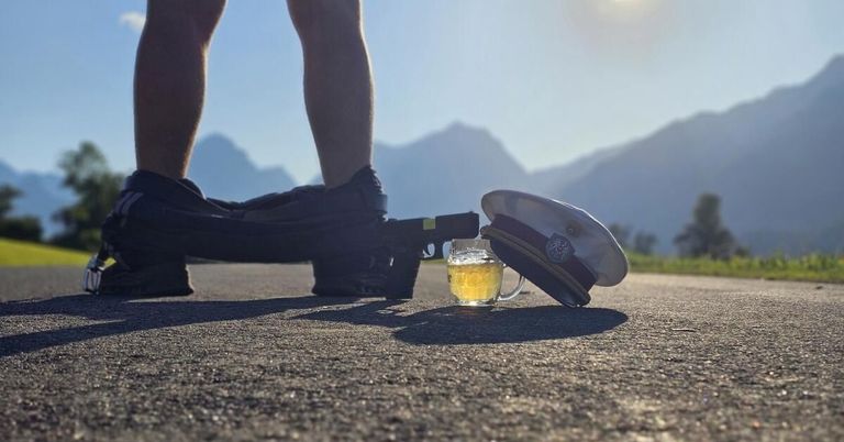 A person's legs in sneakers are shown with a gun, a hat, and a glass of beer on the ground.