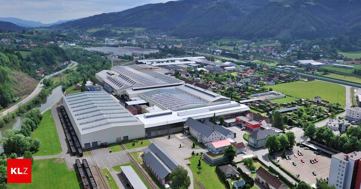 Luftaufnahme eines großen Industriekomplexes mit mehreren Gebäuden, Solarpanelen auf Dächern und einem umliegenden Gebiet mit Häusern, Bäumen und einer Eisenbahnstrecke.