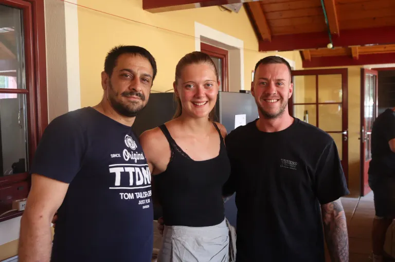 Three people stand close together, smiling for a photo. A man on the left wears a blue shirt with white text, the woman in the middle wears a black tank top, and the man on the right wears a black shirt with white text.