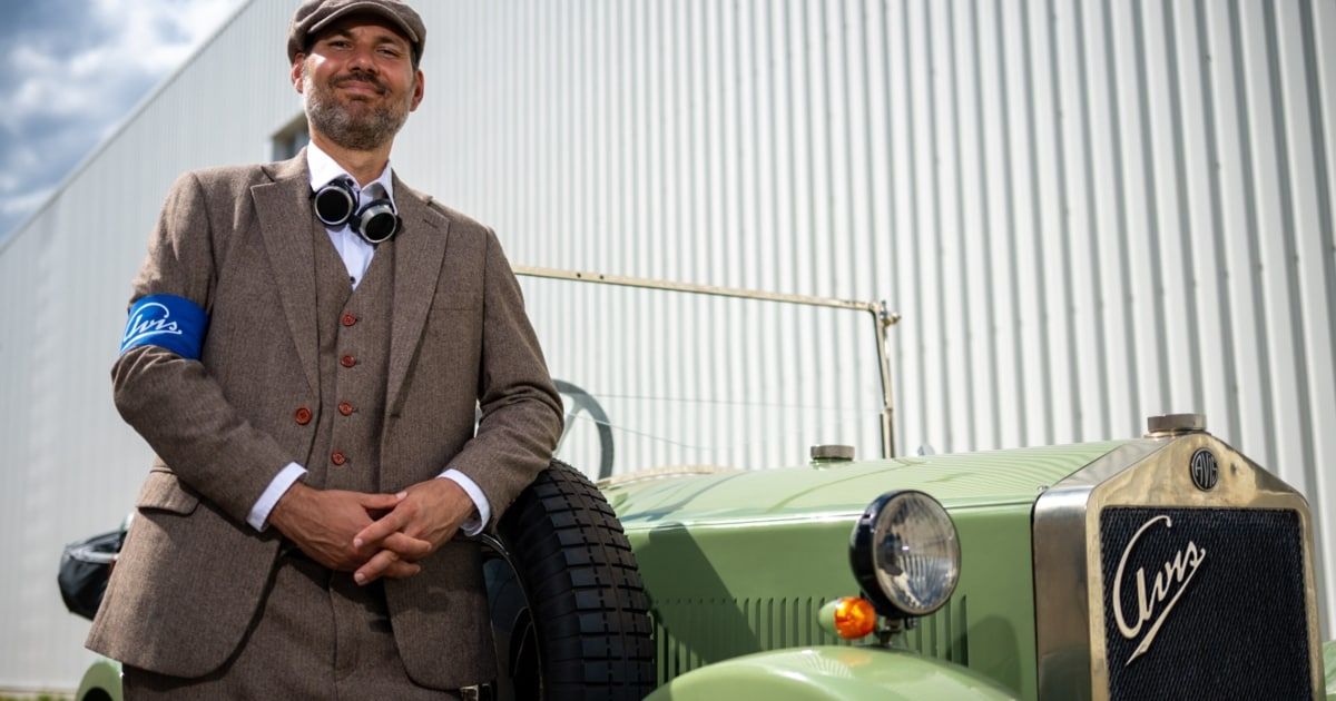 A man in a vintage suit and hat stands next to a green vintage car, smiling and posing for a photo.