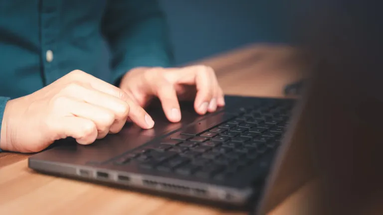 A person's hands are on a laptop keyboard, typing with their fingers. The laptop is on a wooden desk.