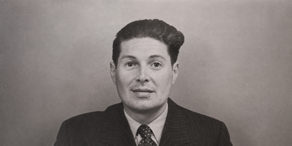 A black-and-white portrait of a man in a suit and tie, with a serious expression and a mole on his right cheek.