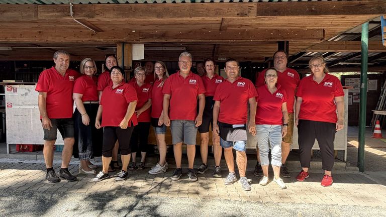 A group of people wearing red shirts with white text stand in a line under a wooden awning. They are smiling and appear to be posing for a photo.