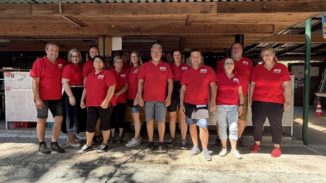 A group of people wearing red shirts with white text stand in a line under a wooden awning. They are smiling and appear to be posing for a photo.