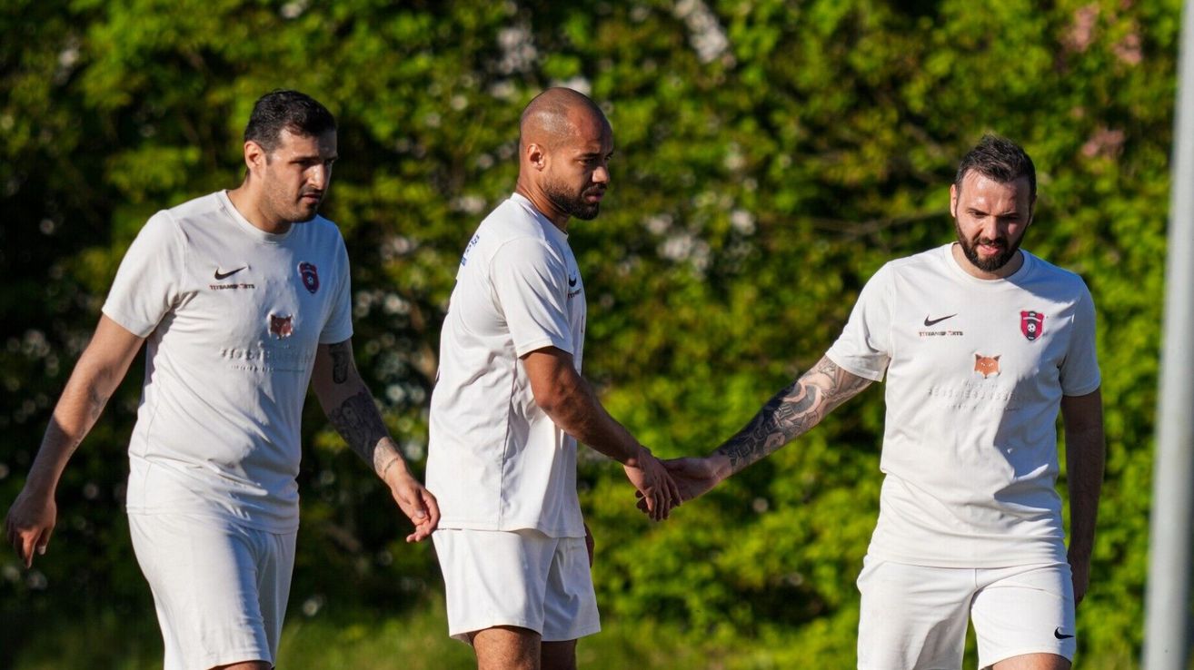 Three men in white uniforms, possibly athletes, are standing in a field, with two of them shaking hands.