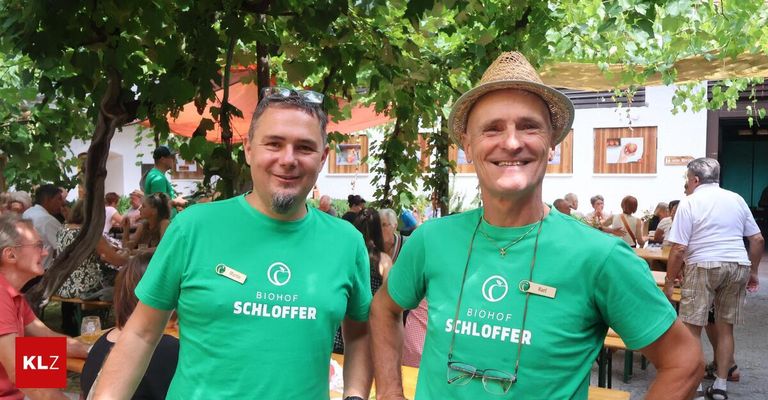 Two men in green t-shirts with the words Biohof Schloffer stand under a tree in an outdoor setting with people sitting and standing behind them.
