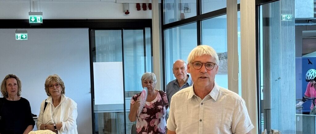 Four people are standing in a room with glass walls and doors. A woman is holding a toothbrush, and a man is looking at her. Another man is standing behind them, and a woman is standing on the left.
